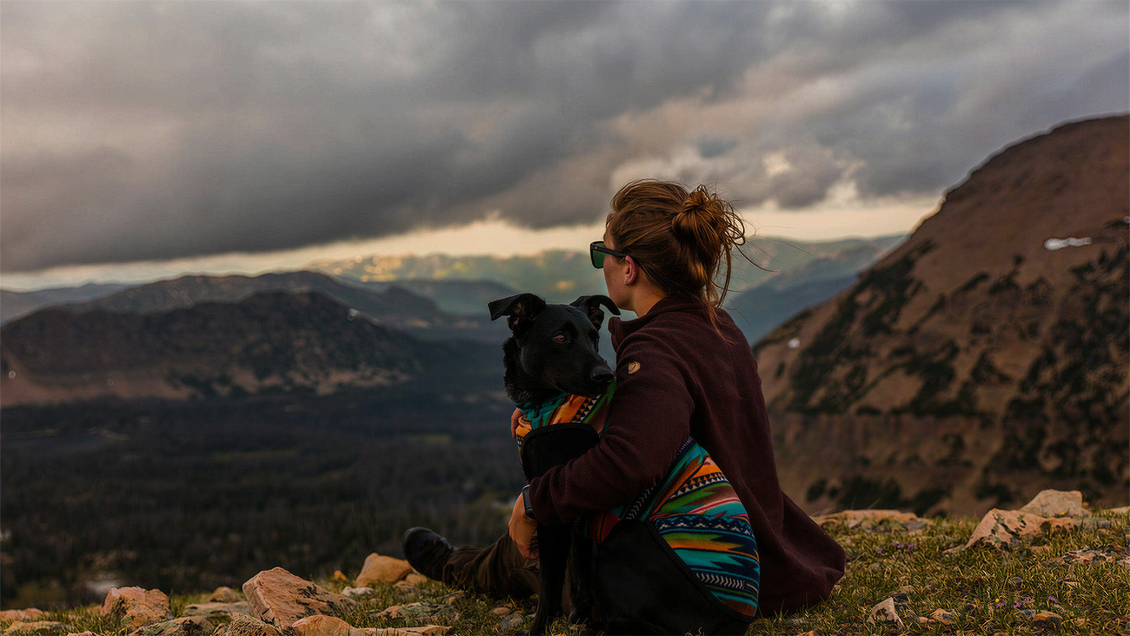 Black dog in a colorful patterned dog apparel being hugged by its owner at the mountain top, staying warm and stylish during outdoor adventures.