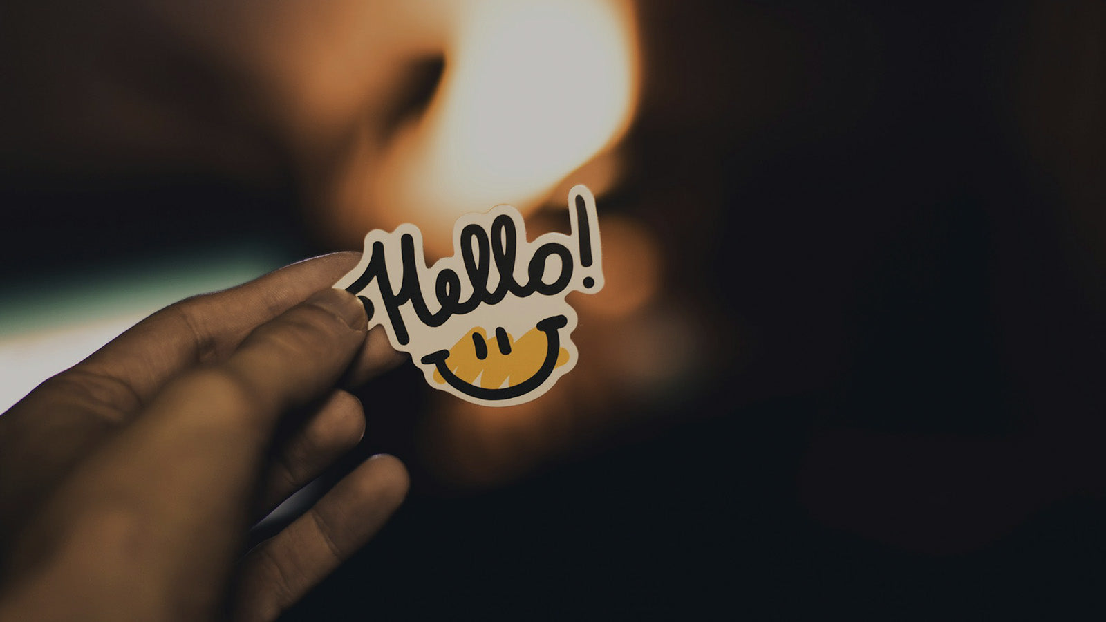 Hand holding a cheerful "Hello!" sticker with a smiley face, symbolizing friendly greetings, positive interactions, and the start of a question-and-answer conversation.