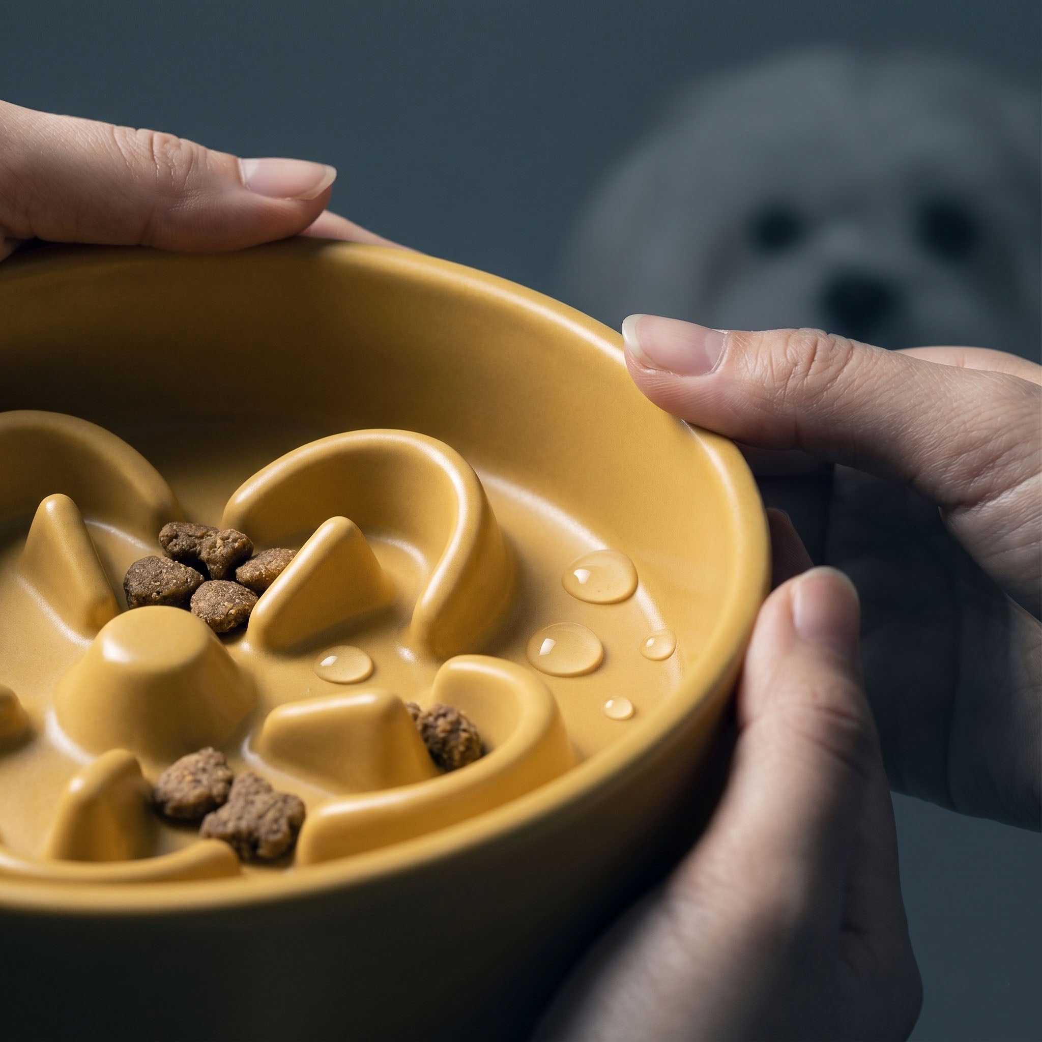 Hands rinsing a yellow ceramic slow feeder dog bowl under water, showing water droplets on the smooth glaze surface.